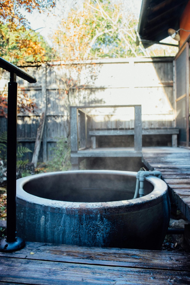 A traditional outdoor wooden onsen tub with steam rising in a Japanese ryokan setting