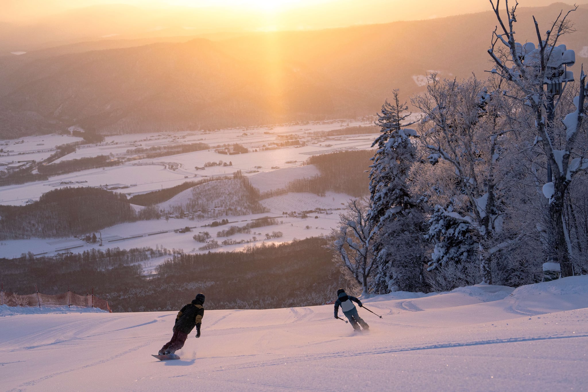 A snowboarder and a skier overlooking the Furano basin from a groomed slope at sunset